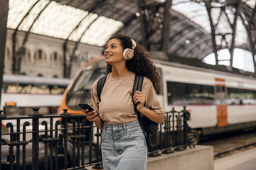 Pretty young african woman with headphones walking through subway station and uses her phone. Brunette wears casual clothes and backpack. Concept digital life, gadgets.