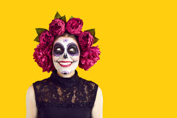 Happy young woman with beautiful spooky sugar skull Catrina makeup isolated on yellow background, looking at copy space side and smiling. Halloween and Day of the Dead aka Dia des los muertos concept