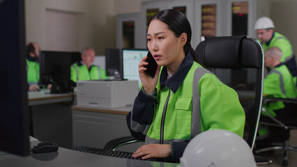 Asian woman technician in reflective uniform talk on phone and work on computer in control room