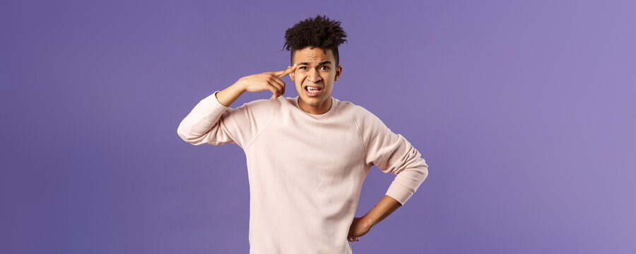 Portrait of angry, annoyed young man scolding someone from being stupid and crazy, rolling index finger over temple staring outraged and irritated camera, standing bothered purple background