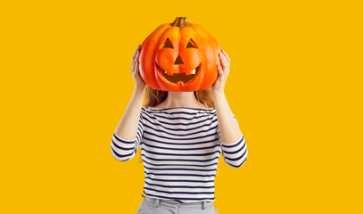 Studio portrait of a woman with a pumpkin head. Funny young girl standing isolated on a yellow background, holding a carved orange pumpkin and hiding her face behind it. Halloween concept - obrazy, fototapety, plakaty
