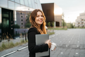 Lifestyle portrait of a young and cheerful woman standing with laptop and coffee cup outdoors. Urban business travel and transportation concept