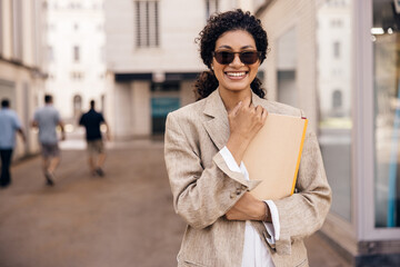 Smiling young african woman looking at camera, posing at europe street with folders. Brunette wears light brown jacket and sunglasses. Concept good mood. © Look!