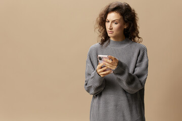 Confused unhappy curly lady in gray casual sweater read conflicting information phone posing isolated on over beige pastel background. Social media, network, distance communication concept. Copy space