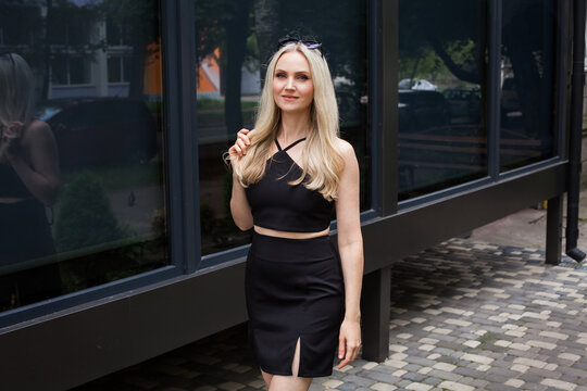 A stylish slender girl with blond long hair and a beautiful smile walks along the city street. Dressed in a short black sexy skirt and top.