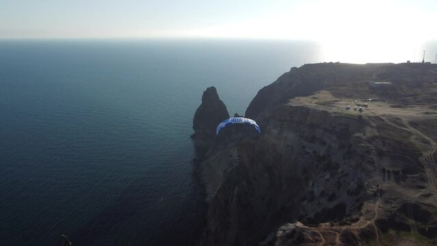 Aerial drone view of a man flying a white and blue paraglider over a hill and trees to the sea waves near the rocks. Active paraglider flight over the seascape with clear skies at suset. Extreme sport