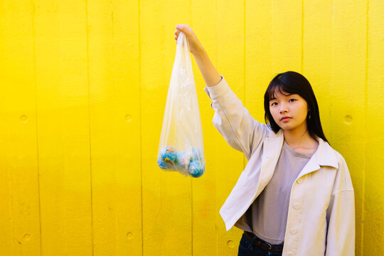 Young woman holding globes in polythene bag with hand raised in front of wall