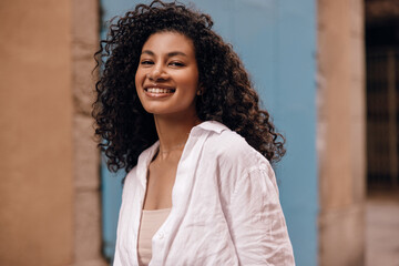 Fototapeta premium Cheerful young african female looking at camera with smile stands on street in spring evening. Brunette hair woman wears singlet and shirt. Concept lifestyle.