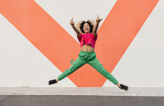 Cheerful woman making obscene hand gesture jumping in front of wall