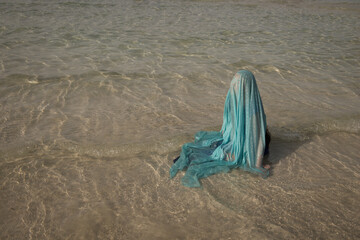 art portrait of girl sitting in sea shore on beach covered by blue thin fabric