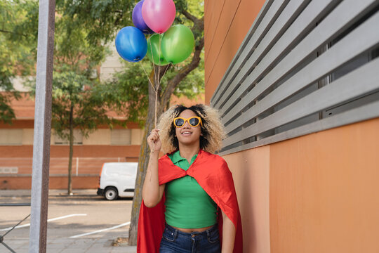 Smiling woman wearing cape standing with multi colored balloons by wall