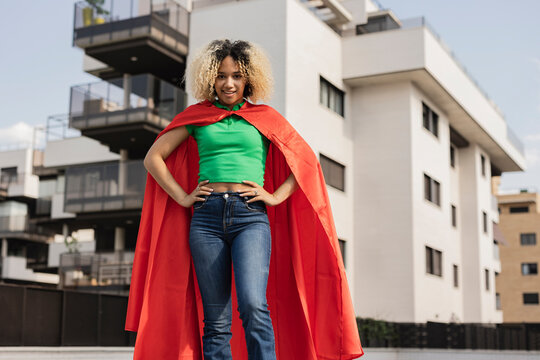 Smiling young woman wearing cape standing with hand on hip in front of wall