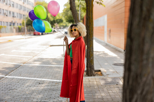 Young woman wearing cape and sunglasses standing with multi colored balloons on footpath