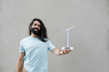 Smiling hipster man with long black hair holding wind turbine model in front of wall