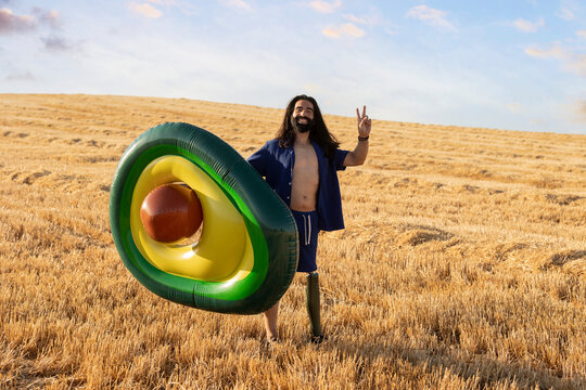 Smiling man with prosthetic leg holding avocado shaped inflatable ring gesturing peace sign at field