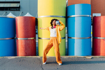 Cheerful young woman carrying boom box on shoulder dancing in front of colorful pipes
