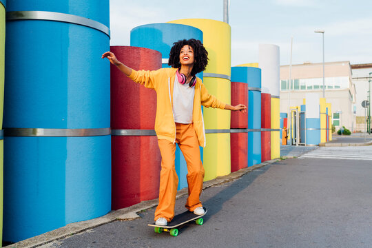 Happy woman with arms outstretched skateboarding by multi colored pipes on sunny day