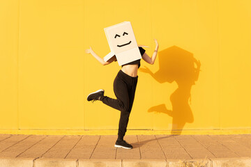 Playful woman wearing box with smiley face gesturing in front of yellow wall on footpath