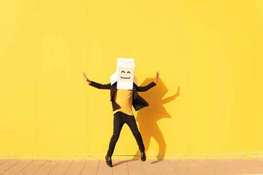 Happy man wearing box with smiley face jumping in front of yellow wall