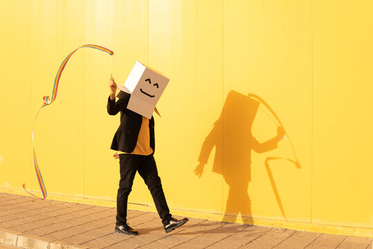 Man wearing box with face spinning rainbow colored rhythmic gymnastics ribbon walking by yellow wall on footpath