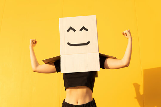 Woman wearing box with face flexing muscle standing in front of of yellow wall