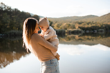 Smiling young woman holding playing with baby boy 1 year old wear knit clothes over nature background and lake with forest. Autumn season. Motherhood.