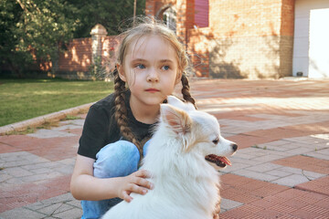 The girl is a child with pigtails, squatting and petting a white fluffy dog of the Spitz breed. Friendship between people and animals. Selective focus
