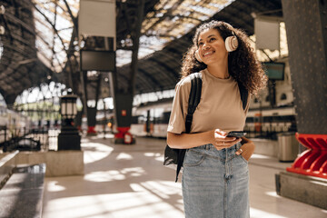 Positive young african woman with overhead headphones looking at digital display, holds smartphone. Brunette with curly hair wears casual clothes and backpack. Concept modern technologies, tourist.