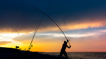 Silhouette of a fisherman casting a rod on the coast at sunset. Night fishing with illuminated alarms.