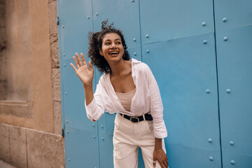 Cheerful young african woman stands on europe street, shaking her hand to say hello to friends. Brunette wears casual clothes. Concept good mood, unexpected encounter.