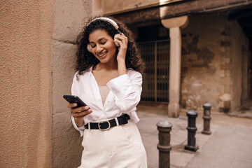Pretty young african girl with overhead headphones stands outdoor in spring day. Model with brunette curly hair wears classic style of clothes. Concept technologies.