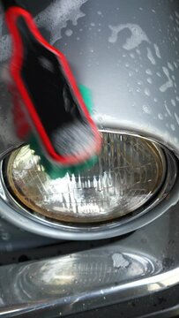 Vertical video social media format – Closeup of a man’s hand using soapy water and a soft brush, to clean a silver wing and a headlamp of an old classic / vintage car.