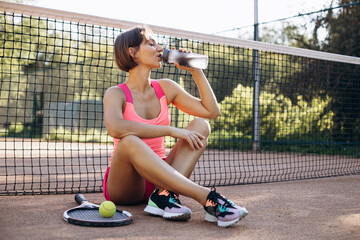 Young woman tennis player sitting on the court and drinking water