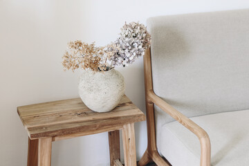 Fall still life photo. Rustic textured vase with dry hydrangea flowers on old wooden bench. Blurred linen mid century sofa background. White wall. Scandinavian interior. Boho elegant home decor.