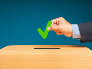 Hand holding a green check mark symbol overhead the voting box at place election with a blue...