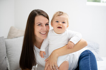 pretty young and happy mother with dark hair, white t-shirt, blue pants is cuddling in bed with her 7 months old son with blue eyes