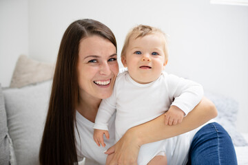 pretty young and happy mother with dark hair, white t-shirt, blue pants is cuddling in bed with her 7 months old son with blue eyes