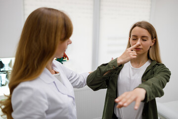 Woman with closed eyes touch nose with fingertip during physical examination. Neurologist checks the coordination of the movements of the female patient