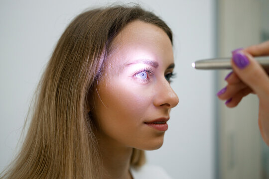 Close up view of doctor testing reflexes of the eye of young woman using a flashlight in medical clinic. Neurological physical examination