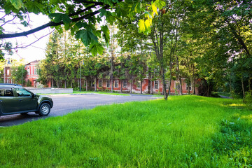 Red brick house among green grass and trees, car on road nearby, summer landscape