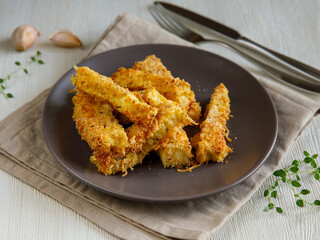 crunchy zucchini sticks breaded with breadcrumbs, parmesan cheese, spices on a black plate with grey napkin on a white wooden table, side view close up, fork and knife, garlic, thyme