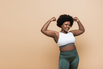 cheerful african american plus size woman in crop top showing power gesture isolated on beige.