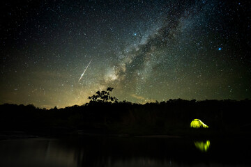 Milky Way and meteor with a yellow tent reflected in the river as a foreground 