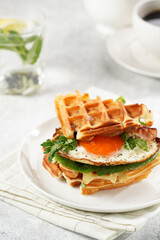 Savory breakfast - fresh homemade belgian waffle sandwich with green lettuce, crisp bacon and fried egg on white plate on beige colored table cloth, glass with lemon water, cup of coffee