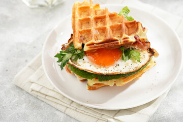 Savory breakfast - fresh homemade belgian waffle sandwich with green lettuce, crisp bacon and fried egg on white plate on beige colored table cloth, glass with lemon water, cup of coffee