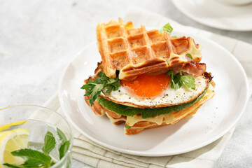 Savory breakfast - fresh homemade belgian waffle sandwich with green lettuce, crisp bacon and fried egg on white plate on beige colored table cloth, glass with lemon water, cup of coffee - obrazy, fototapety, plakaty