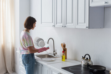 Side view of woman cleaning plate near dishwashing liquid in kitchen.