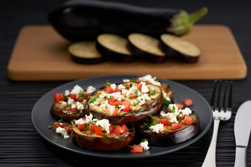 Grilled eggplant slices on a plate, with feta and tomatoes on a black table. whole eggplant background