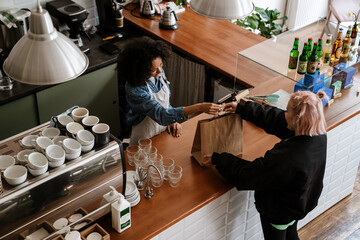 Young black female barista standing by counter with customer in cafe - obrazy, fototapety, plakaty