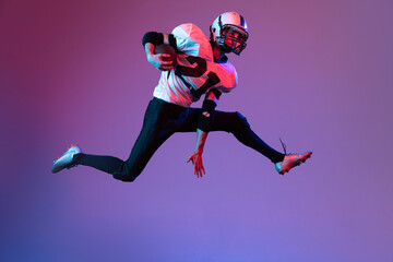 Fototapeta na wymiar Dynamic portrait of sportive man, american football player in uniform playing, training isolated over purple background in neon light.